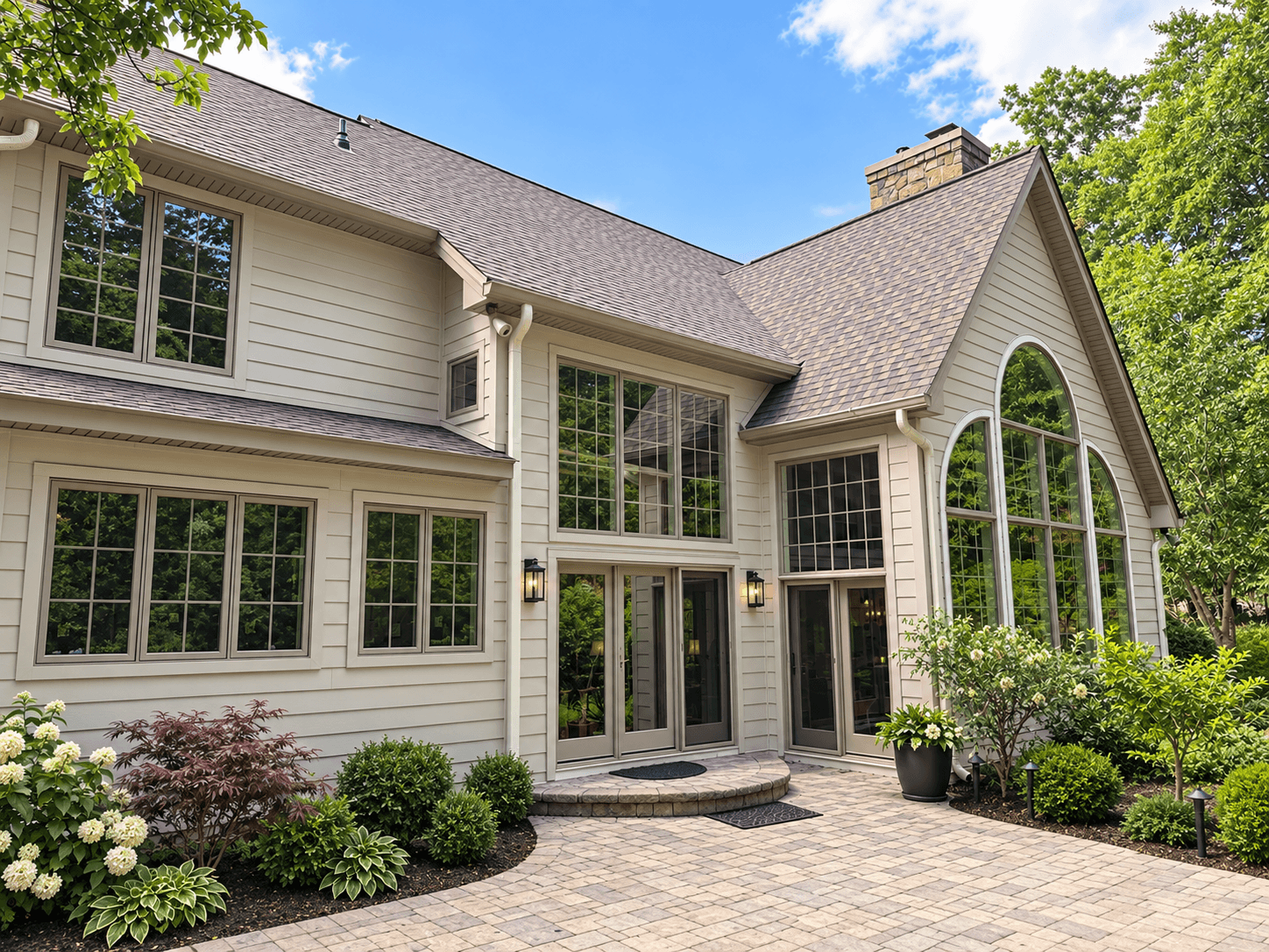Contemporary home exterior with cream siding, gray shingle roof, arched windows, brick chimney, and paved courtyard entry surrounded by landscaped gardens and mature trees