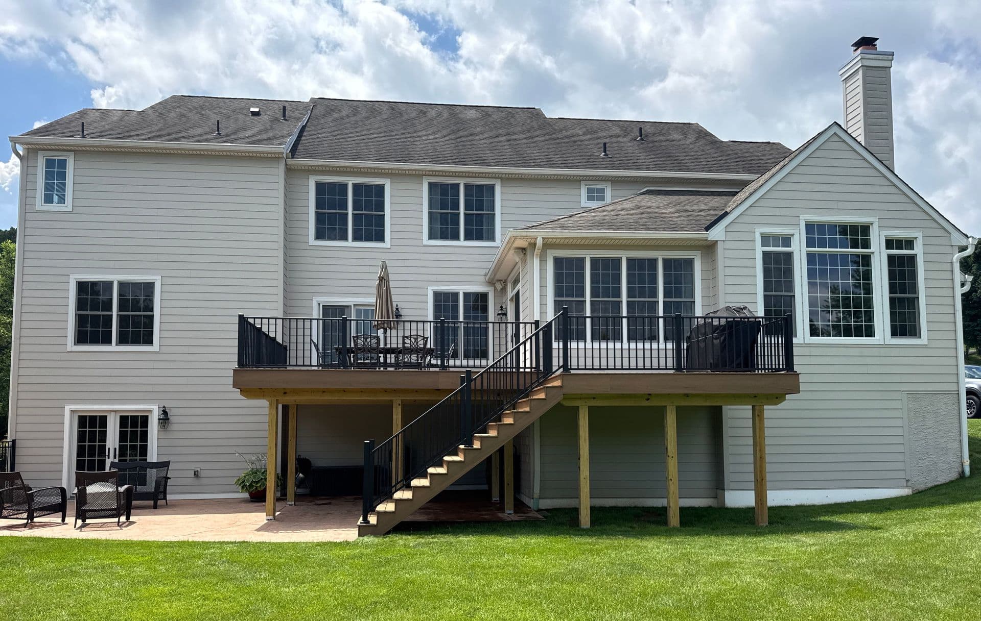 Two-story gray and white residential home with elevated deck, black railings, wooden stairs, large windows, chimney, patio area, and manicured lawn under partly cloudy sky