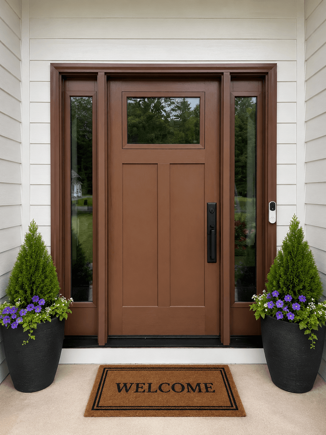 Welcoming front entrance with brown wood door, sidelights, window transom, flanked by planters with purple petunias and green shrubs, welcome doormat, and white siding