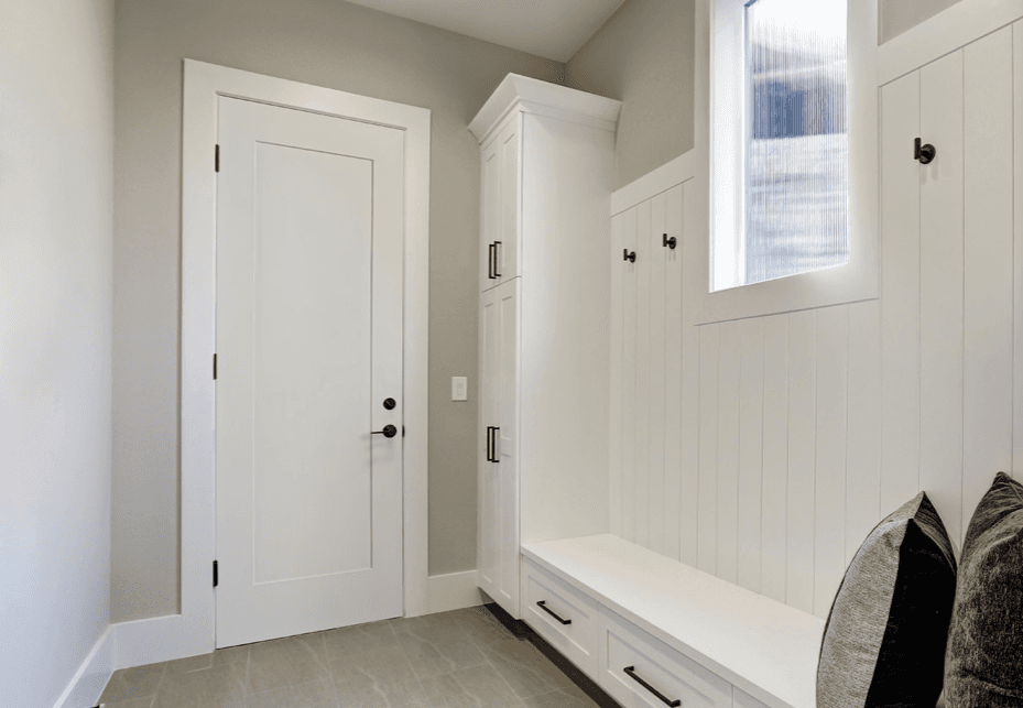 Modern mudroom with white shaker cabinets, built-in bench seating with storage drawers, wall-mounted cubbies with black hardware, framed window with blue shutters, and coats hanging on the right side