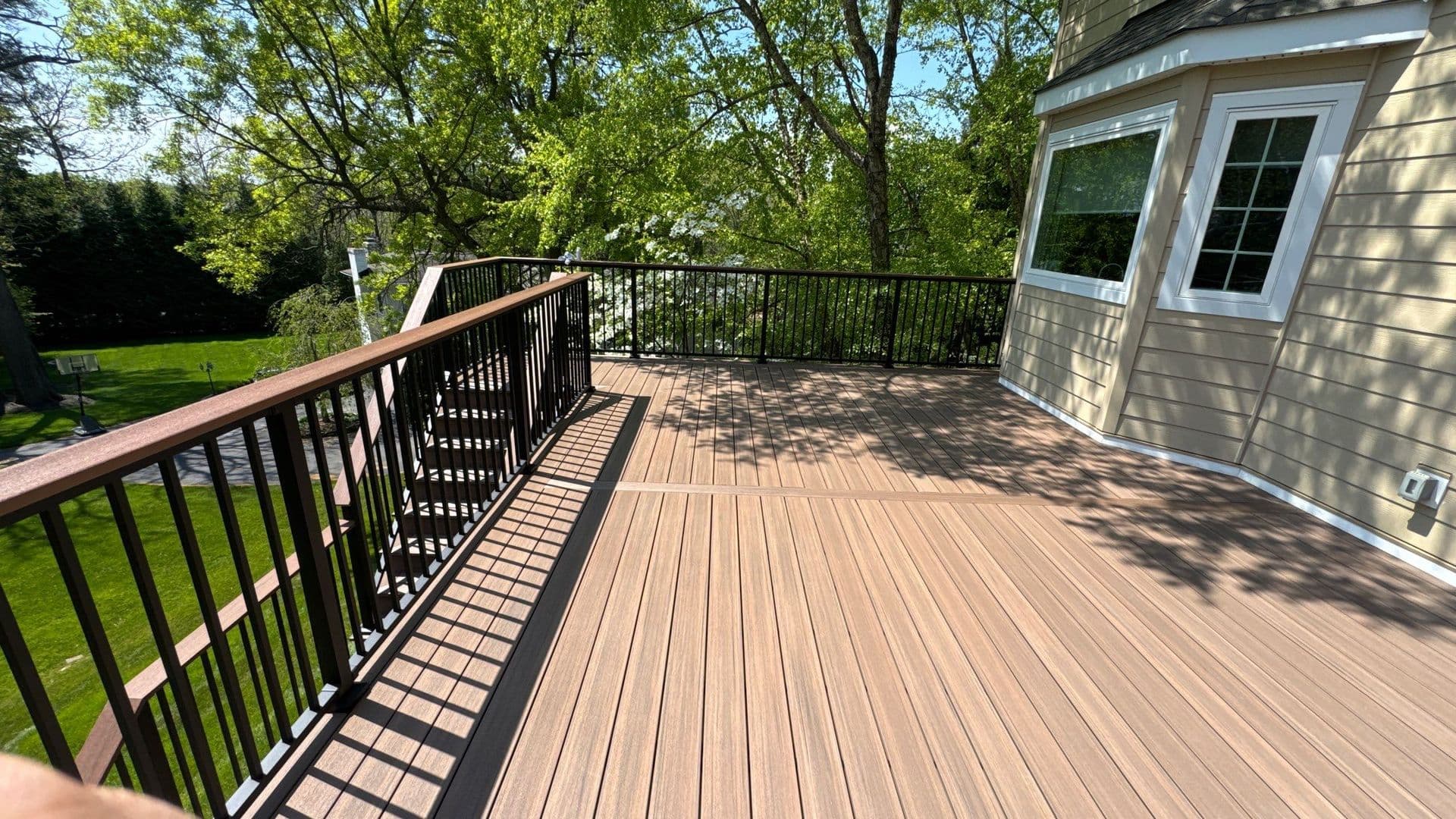 Spacious composite deck with black aluminum railings overlooking a green lawn and mature trees, attached to a house with glass doors and windows