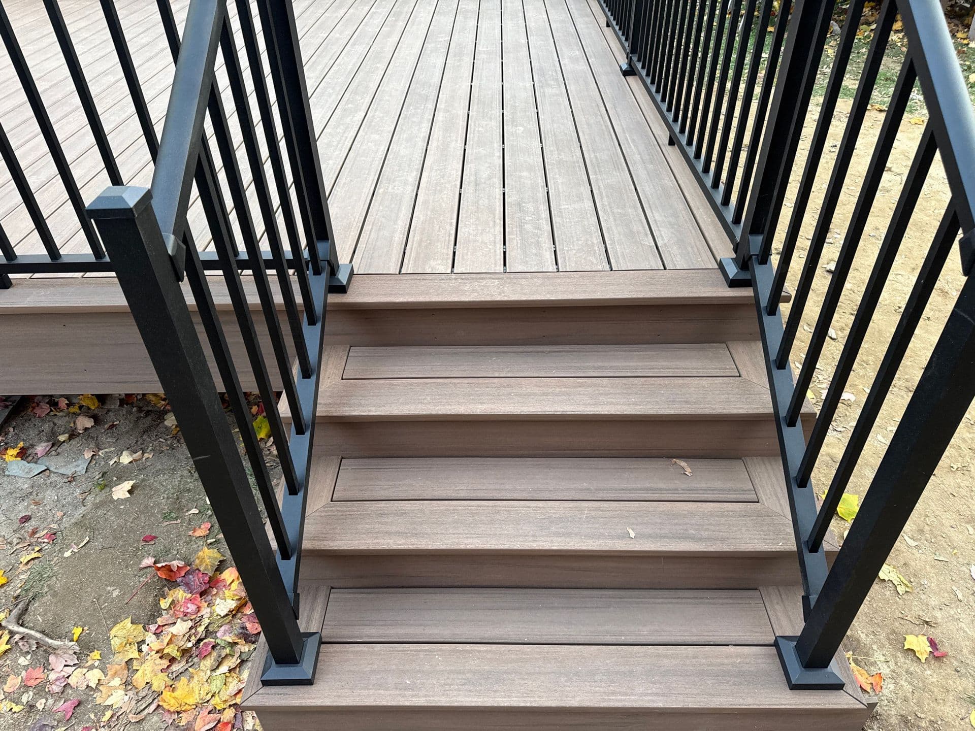 Elevated wooden deck with black railings on both sides, leading to stairs descending to ground level covered with autumn leaves