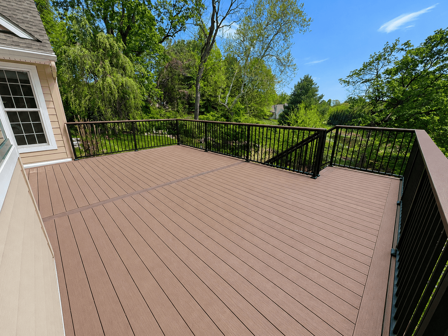 Large composite deck with black metal railing overlooking wooded backyard, attached to house with window visible on left